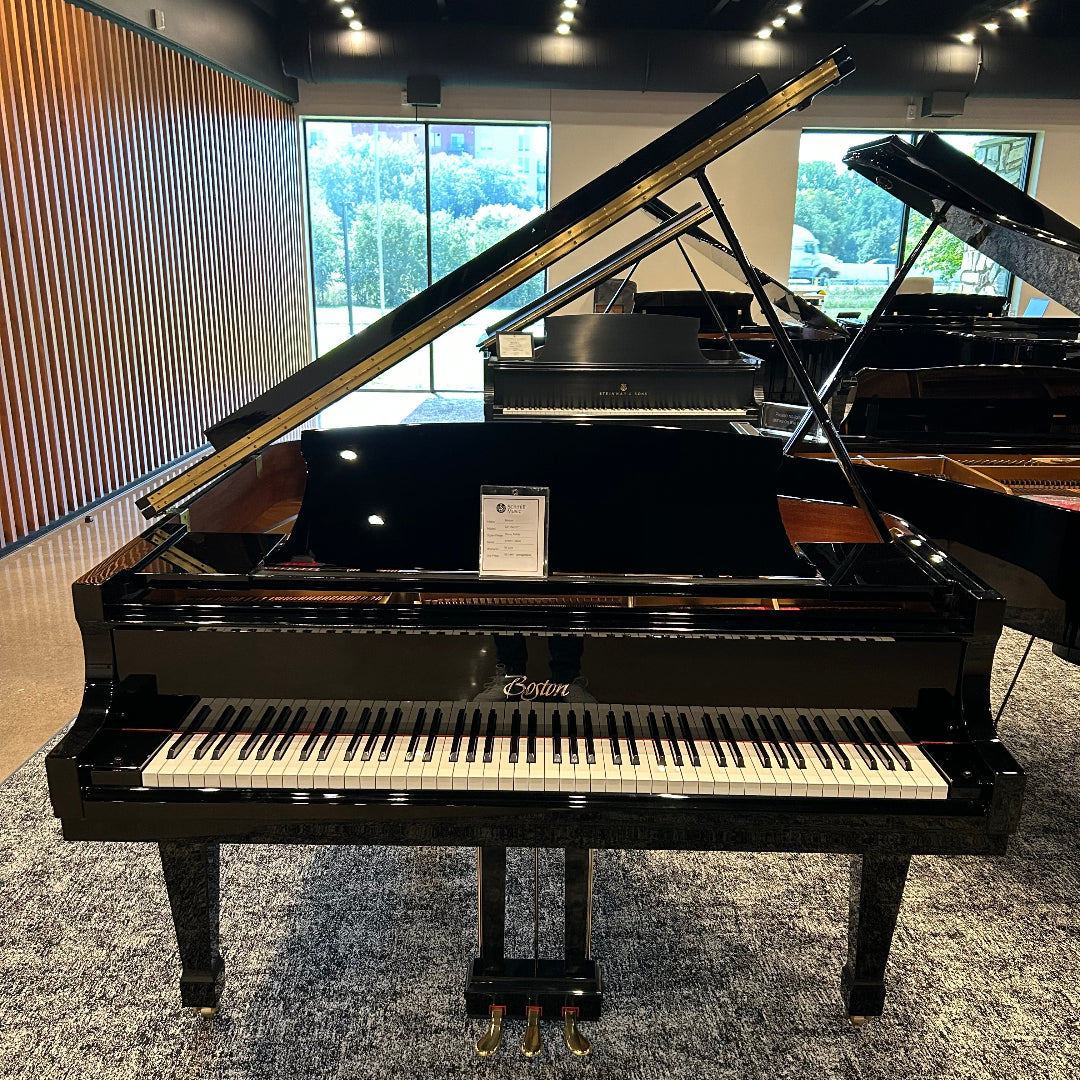 A black grand piano with its lid open, photographed indoors in a showroom with other pianos, windows, and a patterned gray carpet visible.