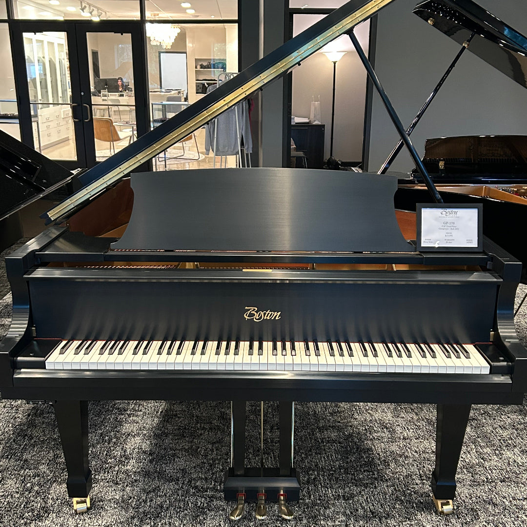 A black Boston grand piano with the lid open, photographed indoors on a gray patterned carpet in a showroom. The Boston logo is visible on the front above the keyboard.
