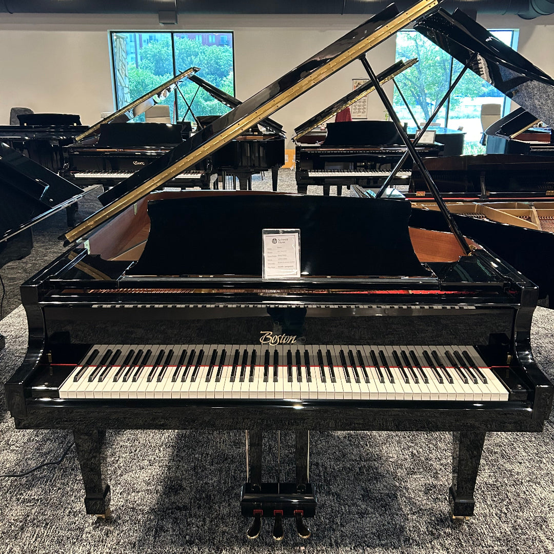 A black grand piano with its lid open, photographed in a showroom with other pianos and windows in the background. The brand name 'Boston' is visible on the front of the instrument.