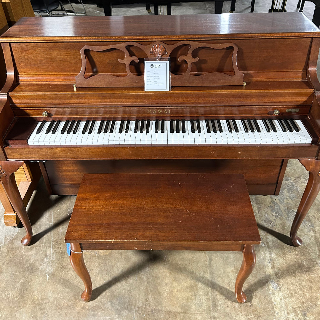 A brown wooden upright piano with a matching wooden bench, photographed in a warehouse or showroom setting with other pianos visible in the background.