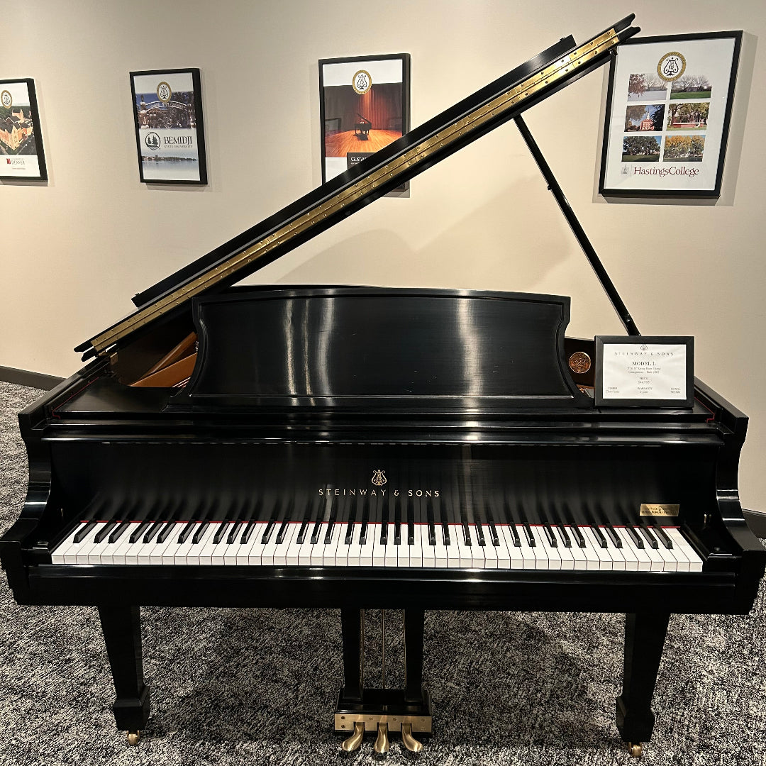 A black grand piano with the lid open, photographed indoors against a beige wall with framed pictures. The brand name Steinway & Sons is visible on the front above the white and black keys.