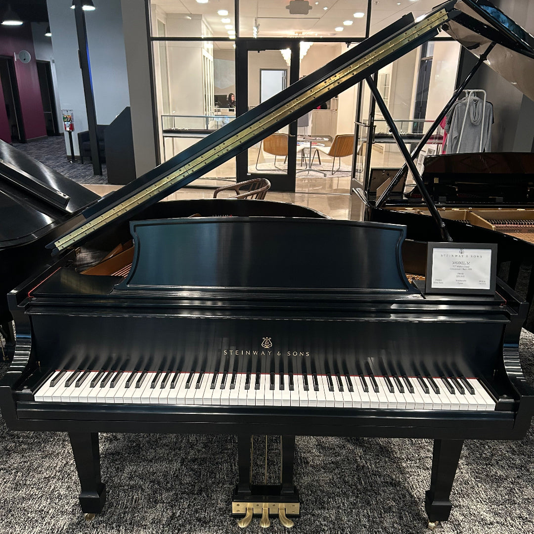 A black Steinway grand piano with the lid open, photographed indoors in a showroom with other instruments and glass windows in the background.