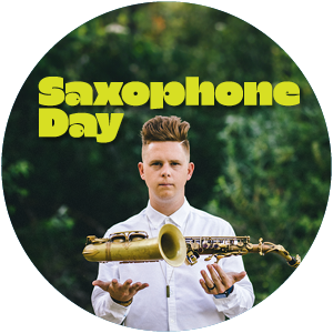 A picture of Alex Hahn, against a backdrop of blurry dark green trees, with his palms facing upward at chest height. An alto Saxophone is hovering slightly above his palms. "Saxophone Day" is above his head in custom green font.  