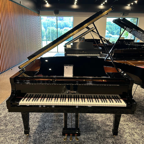 A black grand piano with its lid open, photographed indoors in a showroom with other pianos, windows, and a patterned gray carpet visible.