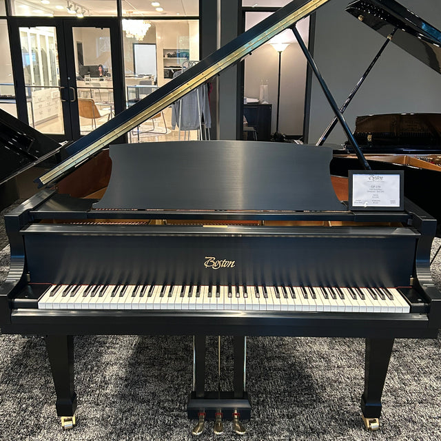 A black Boston grand piano with the lid open, photographed indoors on a gray patterned carpet in a showroom. The Boston logo is visible on the front above the keyboard.