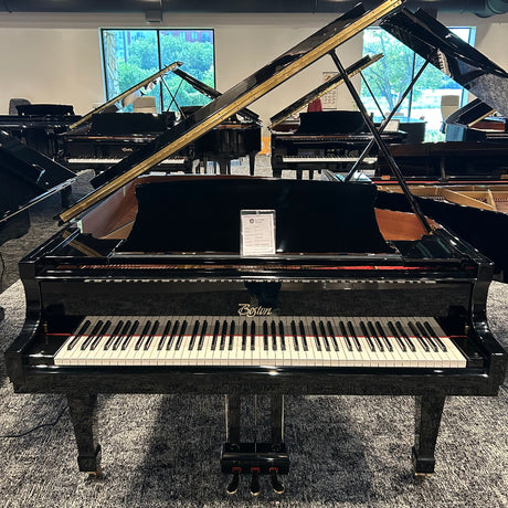 A black grand piano with its lid open, photographed in a showroom with other pianos and windows in the background. The brand name 'Boston' is visible on the front of the instrument.
