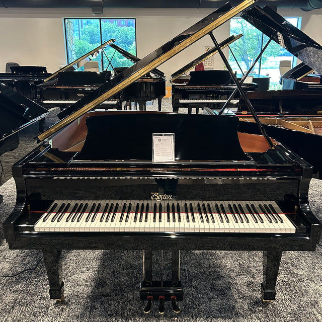 A black grand piano with its lid open, photographed in a showroom with other pianos and windows in the background. The brand name 'Boston' is visible on the front of the instrument.