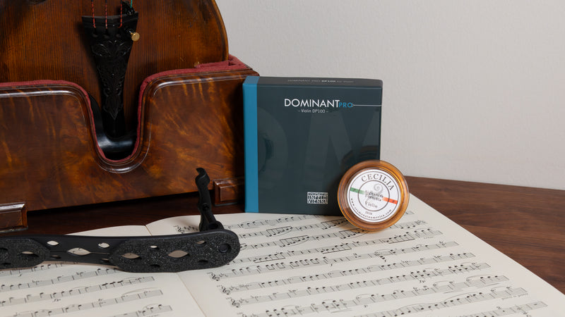 A box of Dominant Tension brand violin strings and rosin resting on sheet music, styled next to a Viola on a wooden stand against a plain light gray wall.