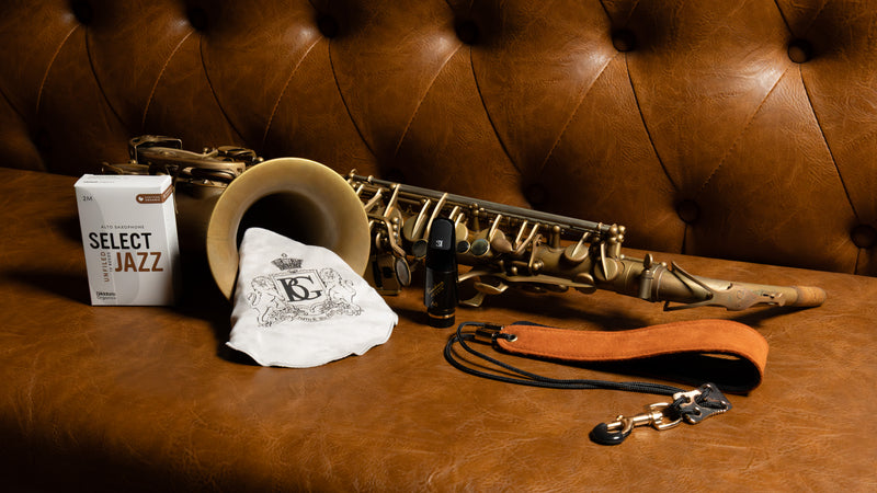 A brass saxophone with a brown leather neck strap, displayed on a brown tufted leather bench alongside a white Select Jazz box, a cleaning swab and a black mouthpiece