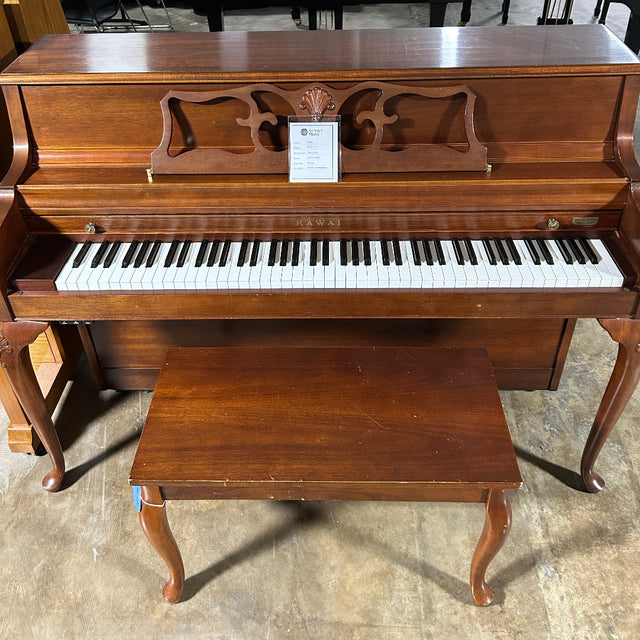 A brown wooden upright piano with a matching wooden bench, photographed in a warehouse or showroom setting with other pianos visible in the background.