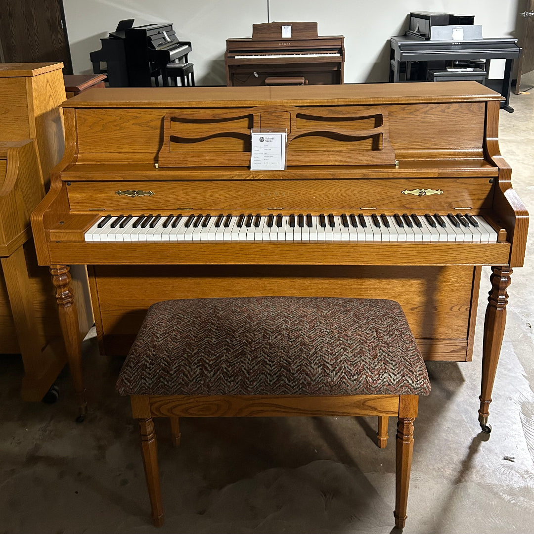 A wooden upright piano with a matching bench, photographed in a showroom or warehouse setting. The piano has a warm medium-brown wood finish, white and black keys, and a patterned brown upholstered bench.