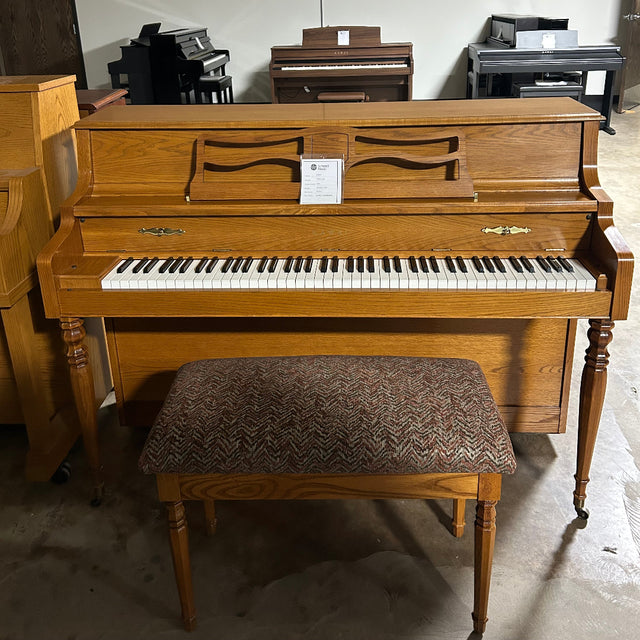 A wooden upright piano with a matching bench, photographed in a showroom or warehouse setting. The piano has a warm medium-brown wood finish, white and black keys, and a patterned brown upholstered bench.