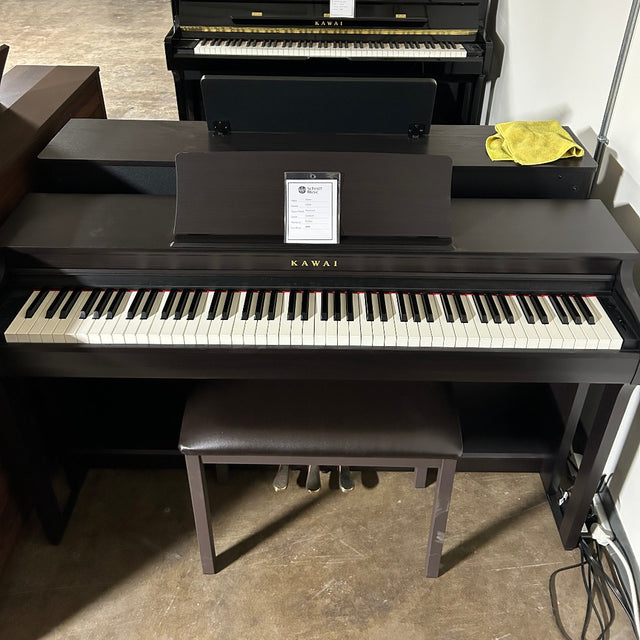 A Yamaha digital piano in dark wood finish with black and white keys, shown in a room setting with a bench and another piano visible in the background.
