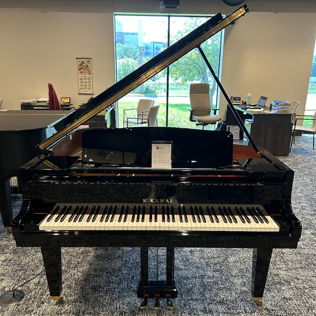 A black grand piano with the lid open, showing the keyboard and interior, photographed in a showroom with windows and chairs in the background.