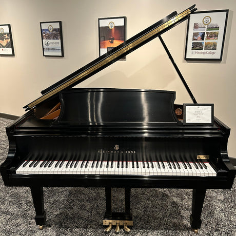 A black grand piano with the lid open, photographed indoors against a beige wall with framed pictures. The brand name Steinway & Sons is visible on the front above the white and black keys.