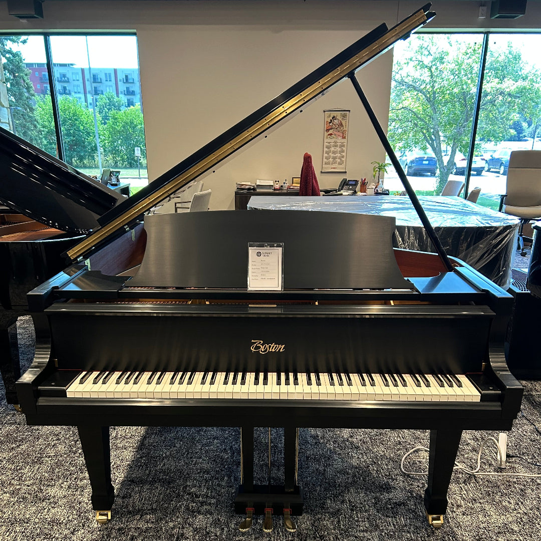 A black Boston grand piano with the lid open, photographed indoors in a showroom with large windows and other pianos visible in the background.