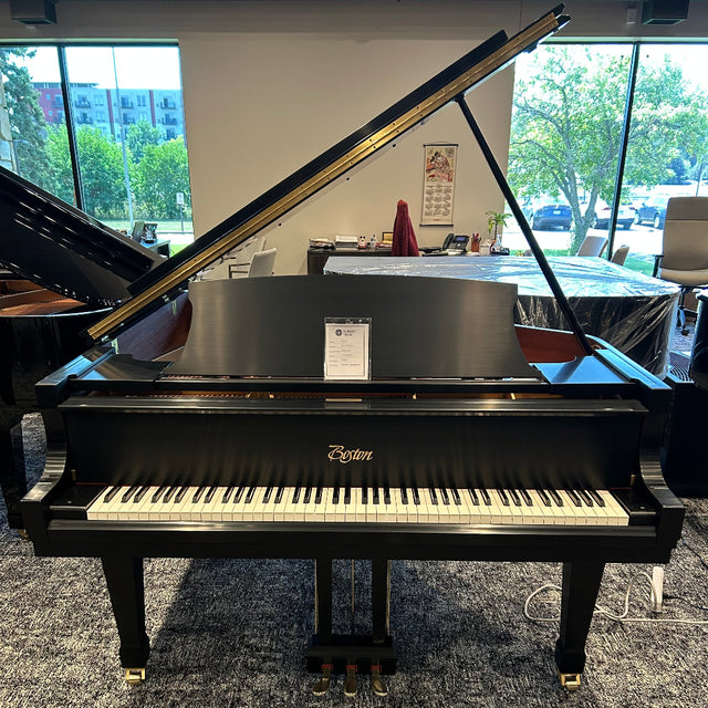 A black Boston grand piano with the lid open, photographed indoors in a showroom with large windows and other pianos visible in the background.