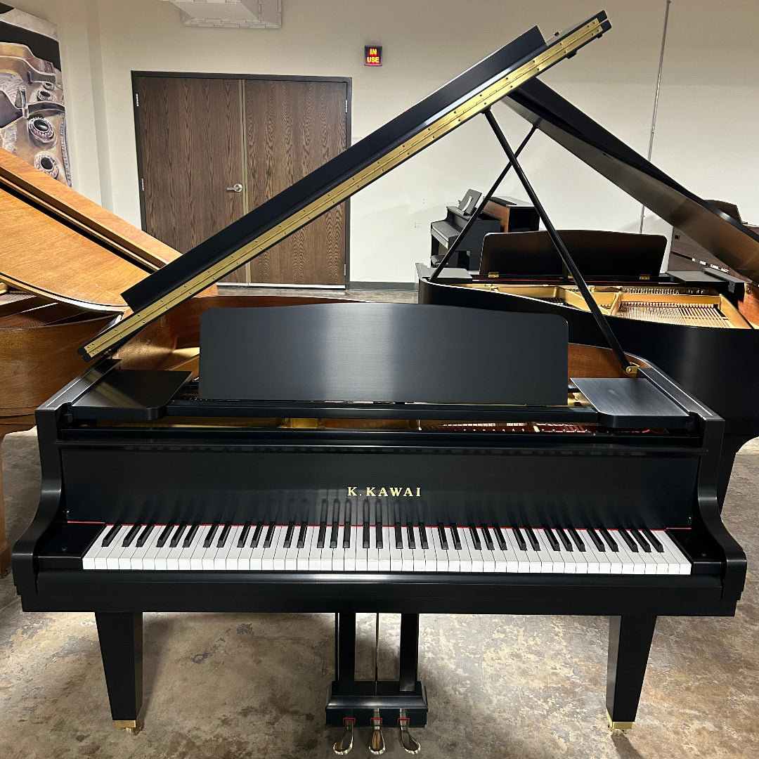 A Kawai grand piano photographed indoors with the lid open, showing the black exterior, white and black keys, and interior wood structure. The piano is placed on a gray concrete floor in a room with white walls and another piano partially visible behind it.