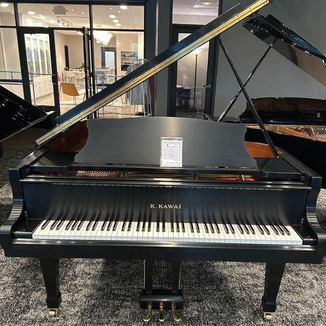 A Kawai grand piano in glossy black, photographed indoors in a showroom with its lid open and keyboard extended. The piano is positioned on a gray patterned carpet, with other instruments and store interior visible in the background.