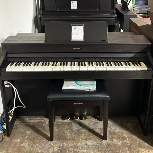 A black digital piano with an attached bench and stand, photographed in a warehouse or showroom setting with shelving and other furniture visible in the background.