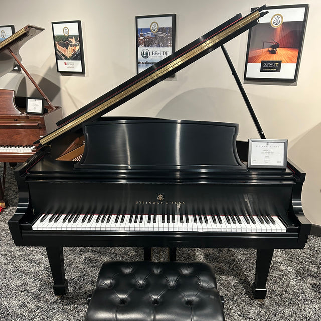 A black grand piano with its lid open, shown with a matching black tufted bench in a showroom setting with framed pictures on the wall.
