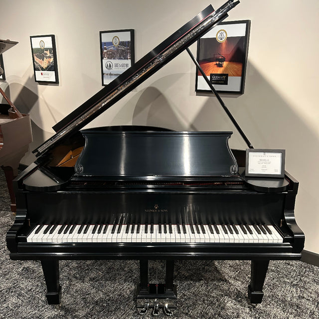 A black grand piano with the lid open, photographed indoors on a gray patterned carpet against a beige wall with framed pictures.