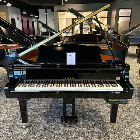 A black grand piano with its lid open, positioned in a showroom with other pianos and musical instruments visible in the background.