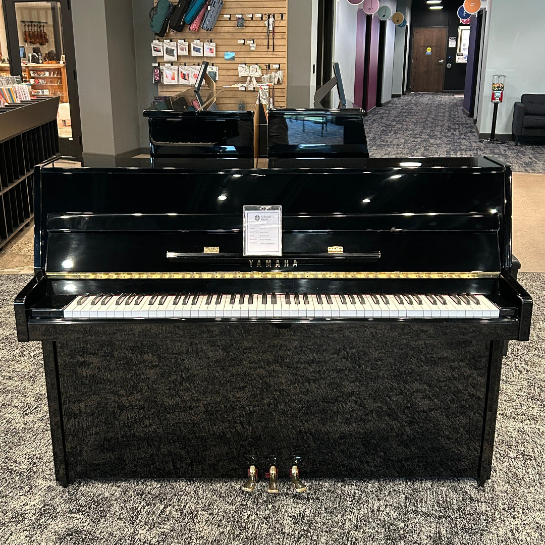 A black upright piano with a glossy finish, shown in a showroom with carpeted floors and display walls in the background.