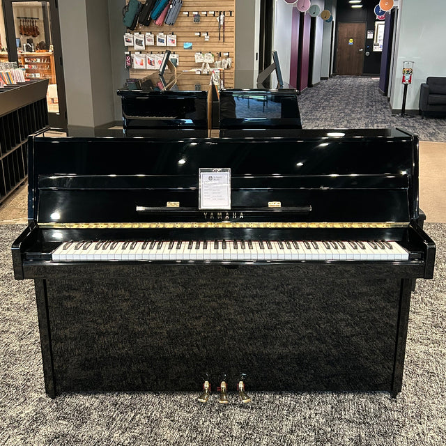 A black upright piano with a glossy finish, shown in a showroom with carpeted floors and display walls in the background.