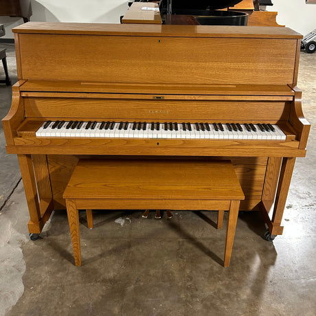 A wooden upright piano with a matching bench and stand, photographed in a warehouse or showroom setting with a concrete floor and white walls.